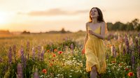 Woman in yellow dress standing in wildflower meadow at golden hour sunset, surrounded by purple lupines and red poppies, holding flowers, looking away thoughtfully in natural outdoor setting.