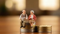 Miniature elderly couple sitting on stack of coins representing retirement planning and financial security for senior citizens in golden light.
