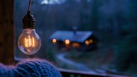 Glowing vintage Edison bulb hanging outdoors at dusk with cozy cabin and warm lights in blurred background, creating atmospheric evening mood with knitted blanket.