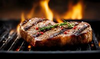 Juicy grilled beef steak with rosemary sprig and peppercorns cooking on flaming barbecue grill, close up view of meat with char marks and fire flames.