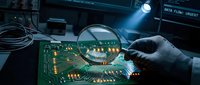 Technician examining computer circuit board with magnifying glass under bright light in electronics repair workshop for quality control and diagnostics.