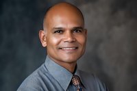 Professional headshot of a mature African American businessman wearing grey shirt and patterned tie, displaying confident smile against dark studio background.