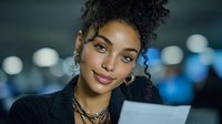 Young African American businesswoman with curly hair reviewing documents in modern office environment with city lights background at night.