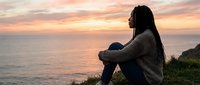 Woman with long dark hair sitting on coastal cliff edge at sunset, wearing casual sweater and jeans, gazing at ocean horizon during golden hour.