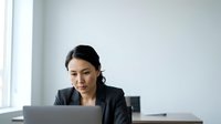 Young woman of East Asian descent, professional businesswoman wearing dark blazer, focused on laptop work at modern office desk with minimalist white background, suitable for corporate and business themes.