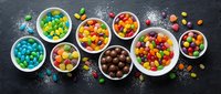 Colorful assorted candies and sweets in white bowls on dark slate background with scattered sugar powder for confectionery and dessert concepts.