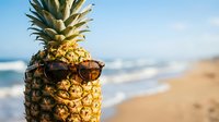 Tropical pineapple wearing sunglasses on sunny beach with ocean waves and sand in background for summer vacation and holiday concept.