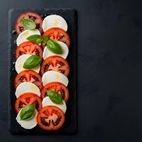 Fresh caprese salad with sliced tomatoes, mozzarella cheese and basil leaves arranged on dark slate board. Classic Italian appetizer for restaurant menus.