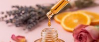 Golden essential oil dripping from a pipette into a glass bottle, with lavender, rose, and orange slices on a pink background.