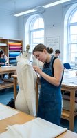 Young Caucasian female fashion design student working on dress form in bright university classroom with fabric shelves and sewing tables.