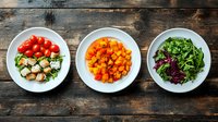 Fresh salad ingredients in white bowls on rustic wooden background, including cherry tomatoes, roasted butternut squash cubes and mixed leafy greens with croutons.
