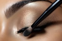 Close-up of makeup application showing professional eyeshadow brush applying shimmer powder to eyelid, demonstrating beauty technique on tan skin tone.