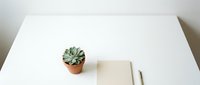 Minimalist white desk workspace with small succulent plant in terracotta pot, blank notebook and pen for productivity and organization.