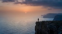 Solitary figure standing on rocky cliff edge overlooking calm ocean at sunset, golden light reflecting on water with distant coastal cliffs visible on horizon.