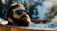 Bearded man with sunglasses relaxing in outdoor hot tub with steam rising, capturing serene wellness moment during spa retreat in natural setting.