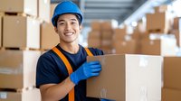 Young Asian male warehouse worker in safety helmet and protective gloves carrying cardboard box in storage facility with stacked packages in background.