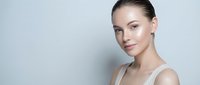 Young Caucasian woman with natural makeup and slicked back hair smiling confidently against neutral background for beauty and skincare campaigns.