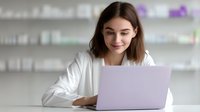 Young female medical professional in white coat working on laptop computer at modern pharmacy or clinic workspace with blurred medicine shelves background.