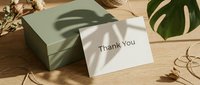 Thank you card leaning against a sage green gift box on a wooden surface with monstera leaf shadow and dried flowers.