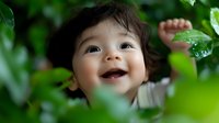 Joyful Asian toddler with bright smile and dark curly hair peeks through green leaves in garden setting, expressing pure happiness and wonder in natural environment.