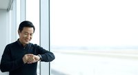 Middle-aged Asian male professional in black shirt checking time on wristwatch by bright window with city view, suitable for business, corporate, and lifestyle content.