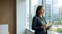 Professional businesswoman holding tablet looking out office window with city skyline view, corporate executive planning strategy in modern workplace.