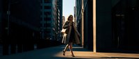 Professional businesswoman walking confidently through urban city street during golden hour sunset with dramatic lighting and modern architecture backdrop.