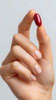 Female hand with manicured nails holding red capsule pill between thumb and index finger against neutral gray background for healthcare or pharmaceutical concept.