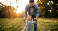 Happy father helping toddler son learn to walk in sunny park during golden hour. Parenting milestone moment with warm natural lighting for family content.