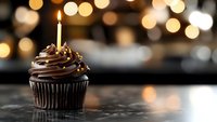 Chocolate cupcake with swirled frosting and lit birthday candle against blurred golden bokeh lights on dark marble surface, creating festive celebration mood.