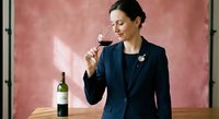 Professional businesswoman in navy suit enjoying wine tasting experience with red wine glass and bottle on wooden table against textured wall background.
