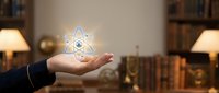 Businessperson holding glowing atom symbol in palm against library background with books and lamps, representing science, innovation, nuclear energy, research and technology concepts.