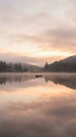 Serene dawn landscape with solitary boat on misty lake surrounded by forested mountains, soft golden sunrise reflecting on calm water surface for nature and travel projects.