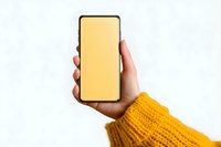 Hand holding smartphone with blank yellow screen in mustard knit sweater sleeve against white background for mockup and app presentation designs.