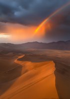 Dramatic rainbow pierces through stormy clouds over vast desert sand dunes at sunset, creating magical light and shadows across rolling orange landscape.