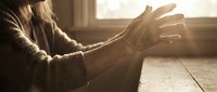 Senior woman with wrinkled hands extended in warm sunlight streaming through window, wearing cozy sweater indoors during golden hour.