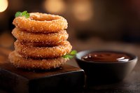 Stack of sugar coated crispy onion rings served on wooden board with chocolate dipping sauce and fresh herbs, close up view in warm lighting.