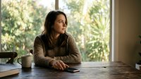 Woman with dark hair and olive complexion, wearing a knit sweater, sits at wooden desk with coffee mug and notebook, gazing thoughtfully out window with natural light and greenery visible behind her, conveying contemplation and focus.