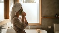 Young woman applying skincare routine in modern bathroom with natural light, wearing towel and robe for morning wellness and self-care ritual.
