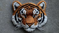 Detailed embroidered tiger head patch with orange, white and black threads on gray fabric background showing intricate needlework and textural details.