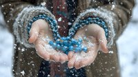 Female hands in knitted mittens holding blue crystal beads in falling snow, creating magical winter atmosphere with soft bokeh effect and blurred background.