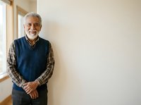 Senior man with gray hair and beard, wearing navy vest over plaid shirt, smiling at camera in bright interior space. Portrait suitable for healthcare, lifestyle, family and senior living concepts.