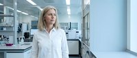Professional blonde Caucasian female scientist in white lab coat standing confidently in modern research laboratory with equipment and clean workspace.