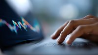 Close-up of trader's hand on laptop keyboard with blurred financial chart in background showing market volatility and stock trading analysis.
