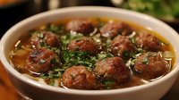 Homemade meatball soup with fresh herbs in clear savory broth served in white ceramic bowl, garnished with chopped parsley and green onions.