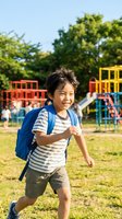 Happy Asian elementary school boy running with blue backpack at colorful playground during sunny day, back to school concept for educational materials.