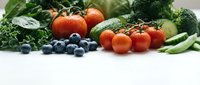Fresh organic vegetables and fruits including tomatoes, broccoli, blueberries and leafy greens arranged on white surface for healthy eating and nutrition.