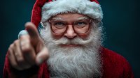 Portrait of senior man with natural white beard wearing red Santa hat and suit, pointing finger at camera with friendly smile against dark background.