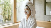 Young Asian woman in white bathrobe and towel turban applying facial mask in modern spa bathroom with bamboo view for skincare and wellness routine.