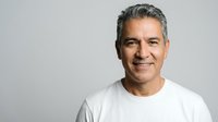 Confident middle-aged Hispanic man with gray hair smiling warmly in white t-shirt against neutral background for professional headshot and lifestyle content.